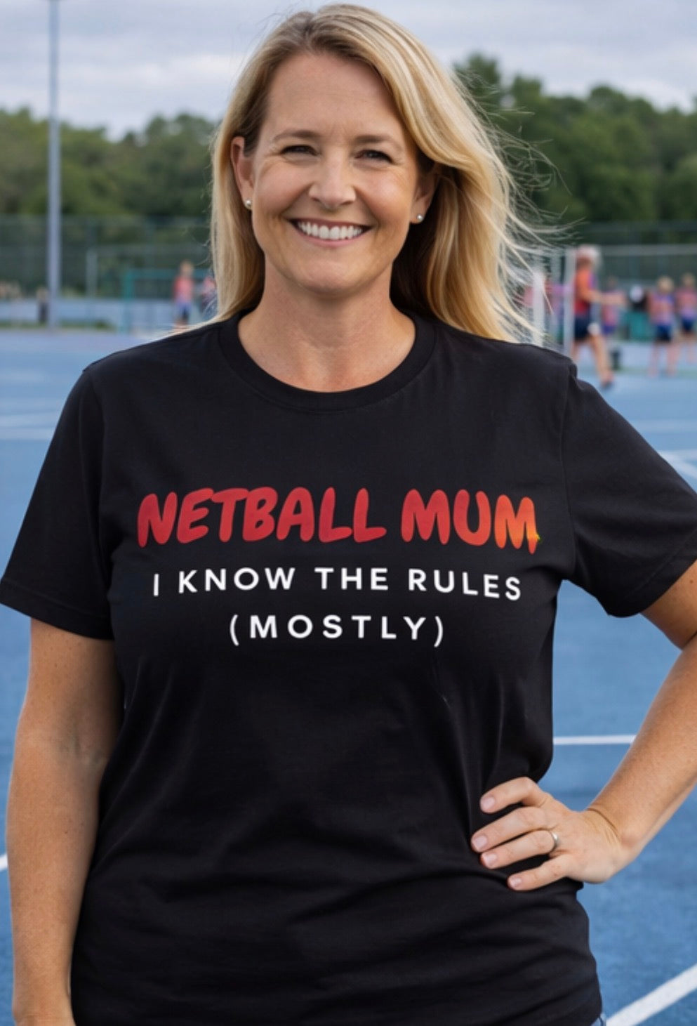 Woman wearing a black t-shirt with 'NETBALL MUM I KNOW THE RULES (MOSTLY)' text on a sports court.