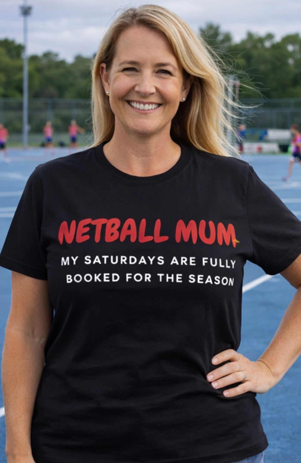 Woman wearing a black t-shirt with 'NETBALL MUM' text on a sports field.