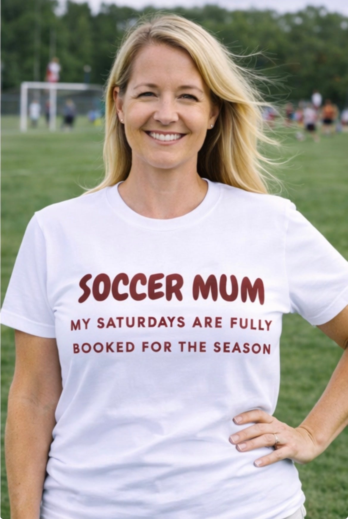 Woman wearing a white t-shirt with 'Soccer Mum' text on a sports field.