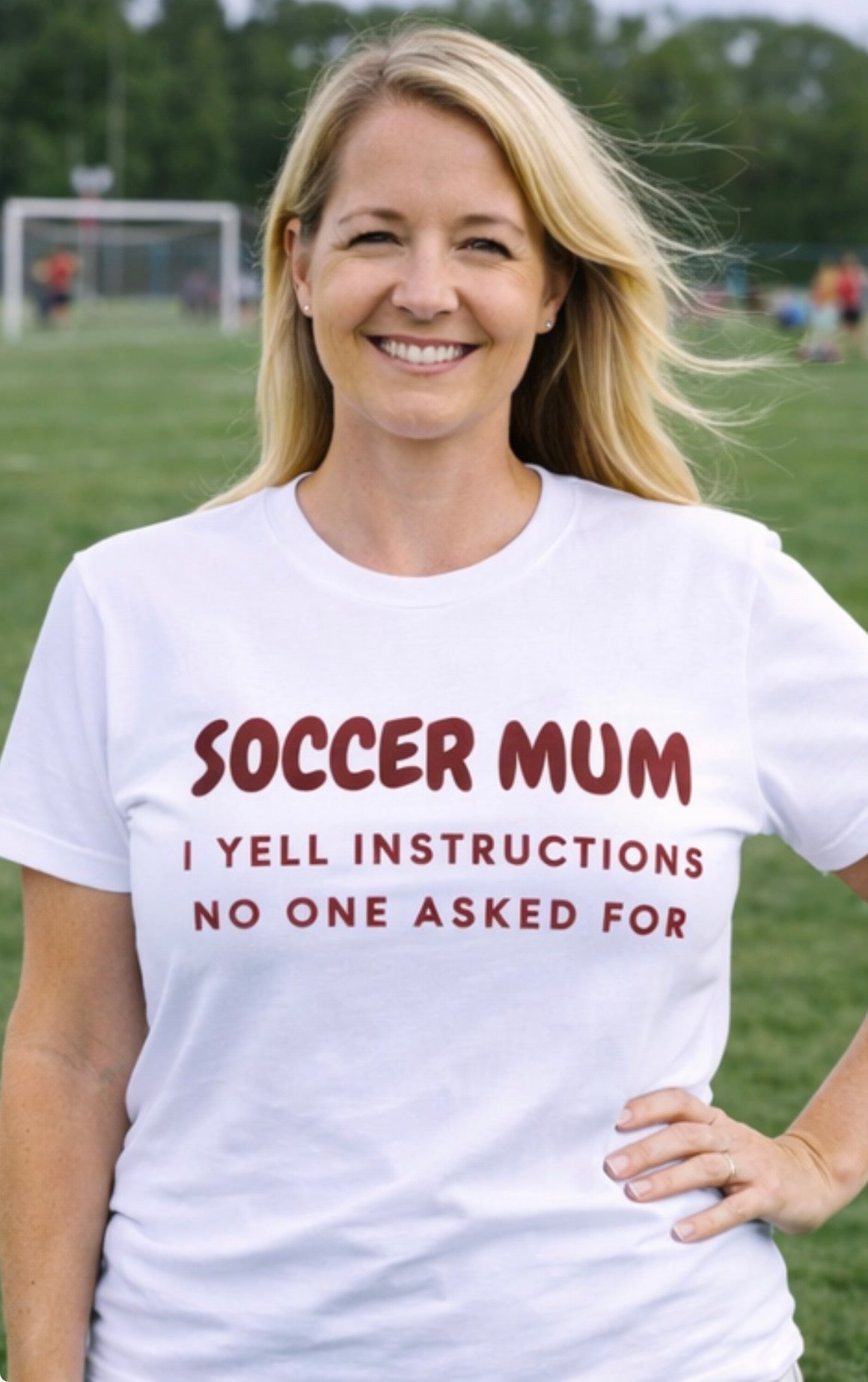 Woman wearing a white t-shirt with 'Soccer Mum I Yell Instructions No One Asked For' text on a soccer field.